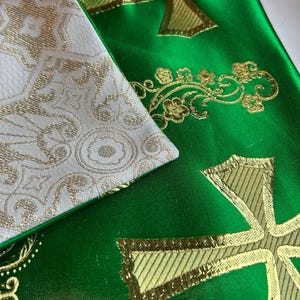 May include: Close-up of two liturgical stoles. One is white with a gold floral pattern, and the other is green with gold embroidered crosses and floral designs. The fabrics have a satin sheen.