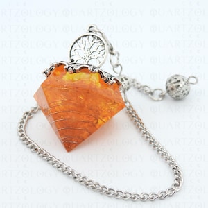 May include: An amber-coloured crystal pendulum with silver-coloured metal detailing. The crystal is faceted and wrapped with wire, suspended from a chain with a tree of life charm and a small ball weight. The background has the word "QUARTZOLOGY" repeated.