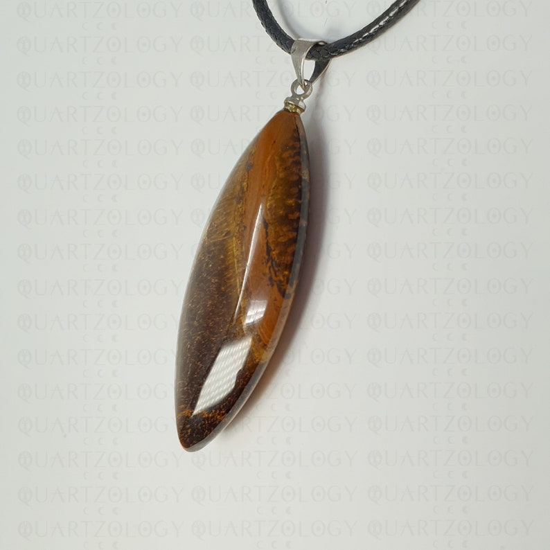 Puede incluir: Un colgante con una piedra preciosa pulida y alargada en tonos marrones y dorados. Cuelga de un cord&oacute;n negro con un cierre met&aacute;lico plateado. La piedra preciosa tiene un acabado liso y brillante. Joyer&iacute;a.