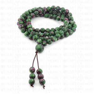 May include: A necklace or prayer beads made from green and maroon gemstone beads, possibly ruby in zoisite. The beads are spherical and of varying sizes. A larger bead and tassel are also present. The background features a repeating pattern of the word "QUARTZOLOGY".