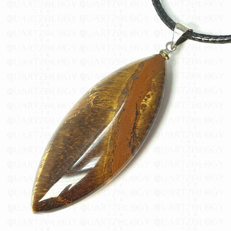 Puede incluir: Colgante alargado de ojo de tigre pulido, con tonos marrones y dorados. Cuelga de un cord&oacute;n de cuero negro con un cierre met&aacute;lico plateado. El colgante mide aproximadamente 7 cm de largo.