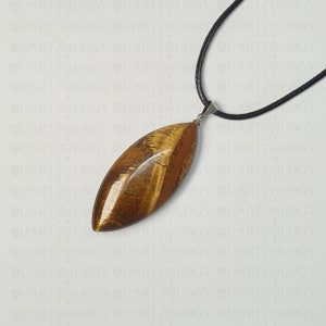 Puede incluir: Un colgante ovalado de ojo de tigre pulido, suspendido de un cord&oacute;n negro. El colgante muestra tonos marrones y dorados, resaltando los patrones naturales de la piedra. Un collar elegante.