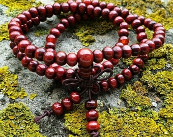 May include: A double strand of dark red wooden beads with a brown tassel. The beads are strung on a cord and are arranged in a circle. The beads are on a mossy green surface.