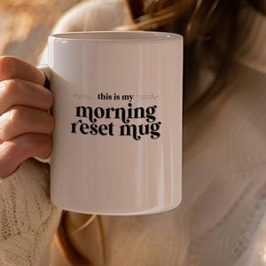 Könnte beinhalten: Eine weiße Keramiktasse mit schwarzem Text, der "this is my morning reset mug" lautet.