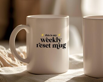 May include: White ceramic mug with a curved handle. The mug features the text "this is my weekly reset mug" in black script with gold star accents. The mug is sitting on a textured surface.