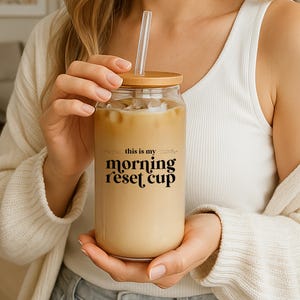Könnte beinhalten: Ein klares Glas mit einem Bambusdeckel und einem durchsichtigen Strohhalm, gefüllt mit einem hellbraunen Getränk. Auf dem Glas steht in schwarzer Schrift "this is my morning reset cup". Das Glas wird von einer Person gehalten.