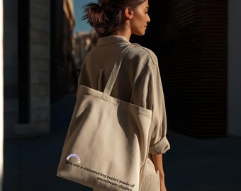May include: A beige canvas tote bag with the text "you are a shimmering comet made of moonbeam giggles." The bag has a purple and white graphic and long handles. The person is wearing a beige outfit.