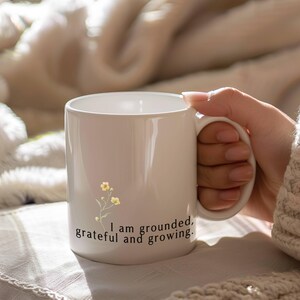 May include: White ceramic mug with a handle, featuring a floral design and the text "I am grounded, grateful and growing." The mug is held by a person. The mug is a standard size for coffee or tea.