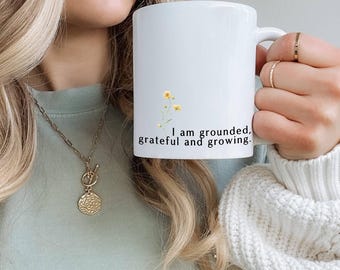 May include: White ceramic mug with a black floral design and the text "I am grounded, grateful and growing."