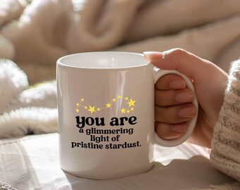 May include: White ceramic mug with a handle, featuring the text "you are a glimmering light of pristine stardust." The design includes yellow stars. The mug is held by a person.