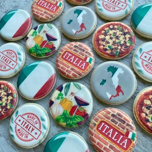 May include: A selection of round, decorated biscuits with Italian designs. Some biscuits show the Italian flag, others depict pizza, maps of Italy, and the text "ITALIA" and "MADE IN ITALY". The biscuits have white icing.