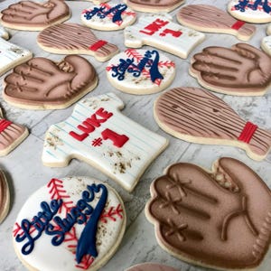 May include: A selection of baseball-themed biscuits, including baseball gloves, bats, and jerseys. The jersey-shaped biscuit has the name "LUKE" and the number "1" in red. Other biscuits feature baseball and baseball bat designs. The biscuits are decorated with brown, red, and blue icing.
