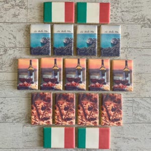 May include: A selection of rectangular magnets with Italian designs. The magnets display images of the Italian flag, coastal scenes with the text "La Bella Vita", wine and glasses, and pizza. The magnets are arranged on a light-coloured surface.