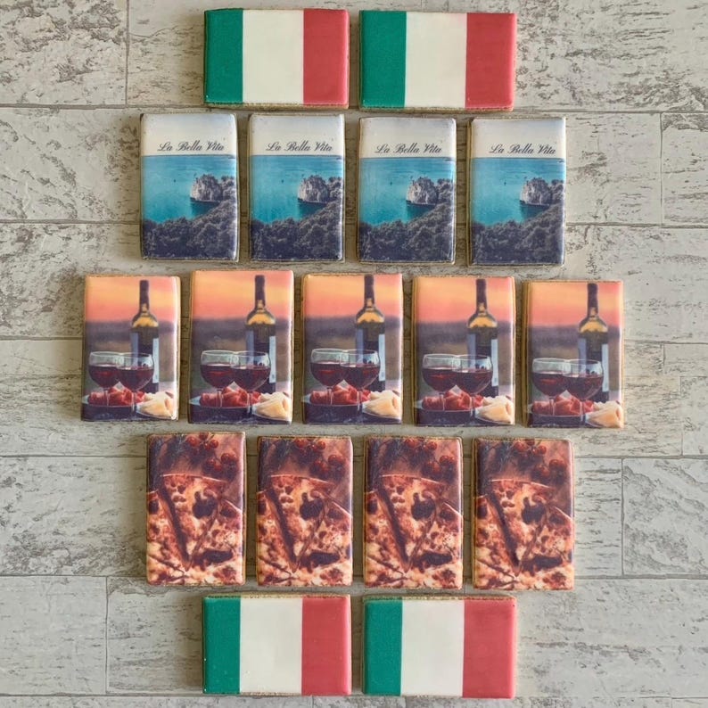 May include: A selection of rectangular magnets with Italian designs. The magnets display images of the Italian flag, coastal scenes with the text "La Bella Vita", wine and glasses, and pizza. The magnets are arranged on a light-coloured surface.