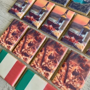 May include: A selection of rectangular biscuits with diverse designs. Some biscuits depict a wine bottle and glasses against a sunset. Others show a pizza design, and a few are decorated with the Italian flag.