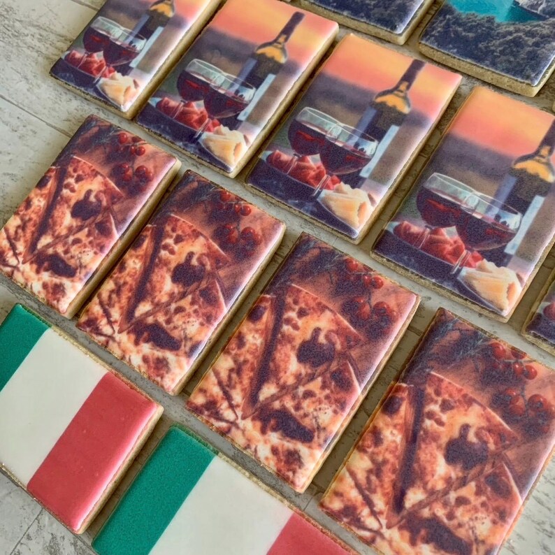 May include: A selection of rectangular biscuits with diverse designs. Some biscuits depict a wine bottle and glasses against a sunset. Others show a pizza design, and a few are decorated with the Italian flag.