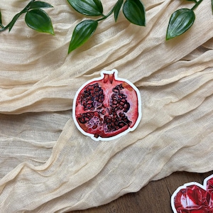May include: A sticker depicting a watercolour illustration of a pomegranate cut in half, showing the red seeds. The sticker is on a beige fabric background with green leaves.