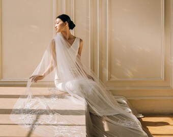 Puede incluir: Una novia con un vestido de novia blanco y un velo se sienta en una estructura de piedra. El vestido tiene un cuello en V y el velo es largo y transparente, con delicados detalles bordados. La escena está bañada por la suave luz del sol.