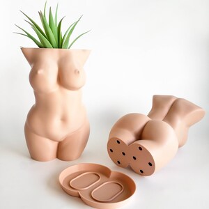 May include: Two nude-colored ceramic planters shaped like the human body. One planter has a plant growing out of the top. The other planter has drainage holes on the bottom. A matching nude-colored ceramic tray is included.