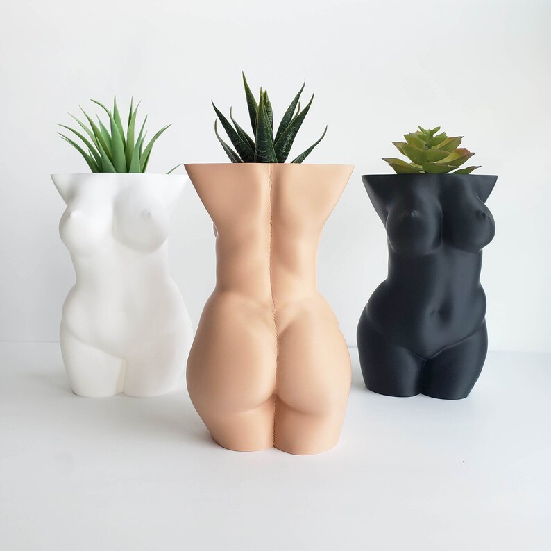 May include: Three ceramic planters shaped like the human body, each with a different color: white, beige, and black. Each planter has a different type of green plant growing inside.
