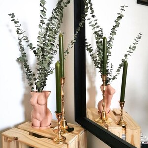 May include: A pair of gold candle holders with green candles, and a nude-colored ceramic vase in the shape of a body, holding a bouquet of dried eucalyptus.