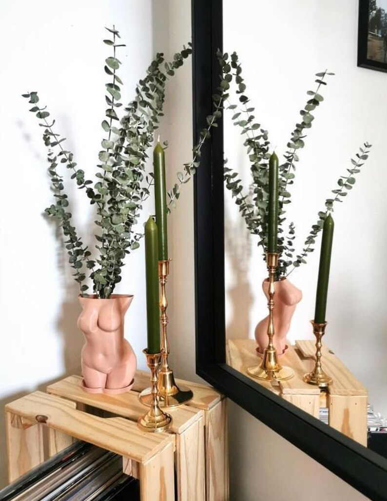 May include: A pair of gold candle holders with green candles, and a nude-colored ceramic vase in the shape of a body, holding a bouquet of dried eucalyptus.