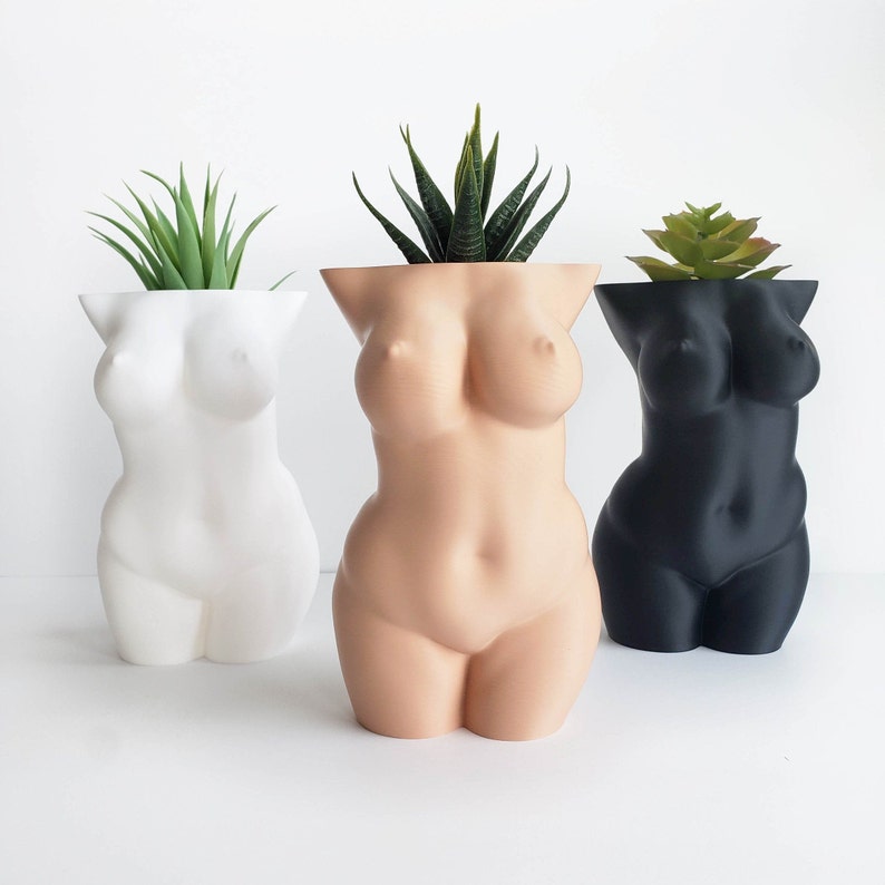 May include: Three ceramic planters shaped like a human torso, each with a different color: white, nude, and black. Each planter has a plant growing out of the top.