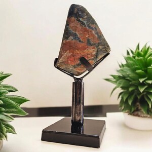 Può includere: Una pietra decorativa esposta su un supporto in metallo nero. La pietra ha una forma triangolare con un mix di colori rosso, marrone e nero. Il supporto ha una base quadrata e un supporto verticale.