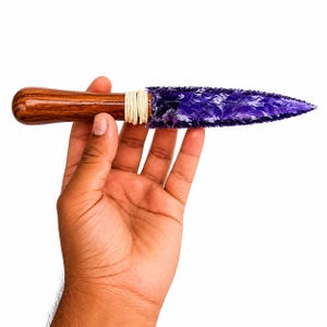 May include: A decorative knife with a wooden handle and a purple, textured blade. The handle is a dark brown color, and the blade is a vibrant purple with a swirling pattern. The handle and blade are connected with a cream-colored twine.