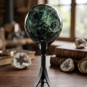 May include: A decorative, dark green crystal ball with a marbled pattern, displayed on a black metal stand. The sphere is set against a backdrop of a wooden table, books, and natural light, creating a sophisticated, elegant aesthetic.