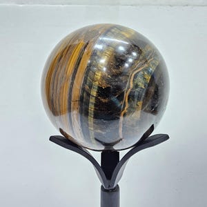May include: A polished tiger's eye sphere, showcasing bands of gold, brown, and blue hues. The gemstone is mounted on a black metal stand with a stylized floral design, creating a striking display piece.