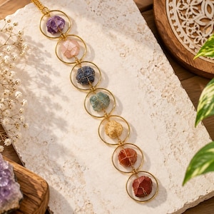 May include: A vertical wall hanging ornament featuring seven gold-toned metal rings connected in a chain. Each ring contains a single raw, wire-wrapped gemstone in varying colours including purple, pink, dark blue, teal, yellow, orange, and red.