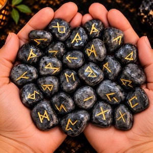 Pode incluir: Uma coleção de pedras polidas cinza-escuras com texturas salpicadas, cada uma gravada com um símbolo rúnico dourado único. As pedras são lisas, arredondadas e variam ligeiramente na forma, apresentando caracteres gravados preenchidos com tinta dourada.
