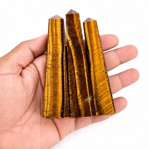 Op de afbeelding: Drie gepolijste tijgeroog kristaltorens met een puntige bovenkant en een zeshoekige basis. De stenen hebben een chatoyante goudbruine kleur met donkerbruine verticale banden en een gladde, reflecterende textuur over de gehele oppervlakte.