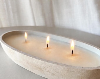 May include: An oval-shaped, light beige concrete candle vessel containing off-white wax with three lit wicks. The container has a textured, matte finish and a shallow depth, holding the wax evenly across the entire interior surface of the bowl.