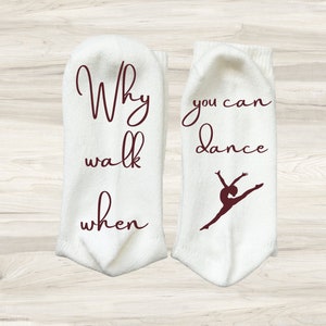 Puede incluir: Un par de calcetines blancos con texto marrón. Un calcetín dice "¿Por qué caminar cuando puedes bailar?" y el otro calcetín tiene una silueta de una bailarina.