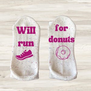 Könnte beinhalten: Ein Paar weiße Knöchelsocken mit gesprenkeltem Muster. Eine Socke hat die Worte "Will run" mit einer Schuhgrafik, die andere "for donuts" mit einer Donutgrafik. Text und Grafiken sind magenta.