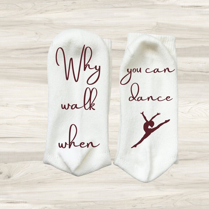 May include: A pair of white socks with maroon text. One sock says "Why walk when you can dance" and the other sock has a silhouette of a dancer.