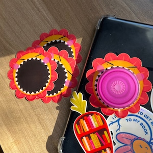 May include: Three red and yellow flower stickers with a black centre and white teeth. The stickers are on a wooden surface. There is a phone case with a sticker of a carrot with books on a shelf, a pink circle, and the text "To get to my book".