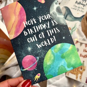 May include: A birthday card with a space theme. The card shows planets, stars, and the text "Hope your birthday is out of this world!" in white. The planets are in shades of pink, green, and grey.