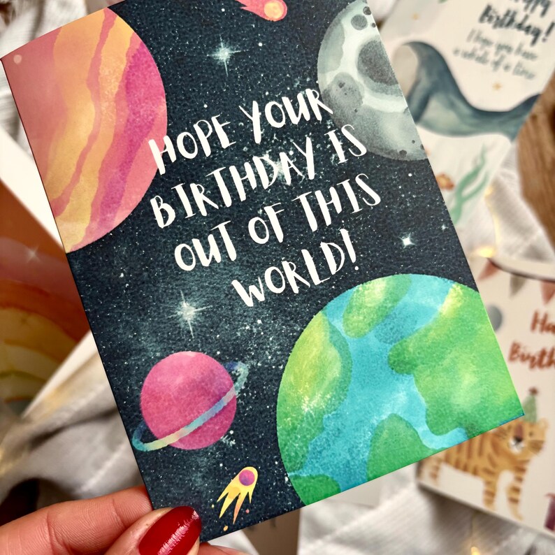 May include: A birthday card with a space theme. The card shows planets, stars, and the text "Hope your birthday is out of this world!" in white. The planets are in shades of pink, green, and grey.