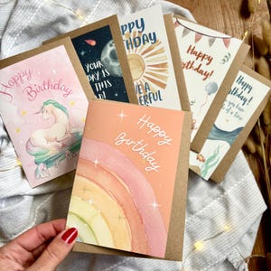 May include: A selection of birthday cards with different illustrations and messages. Designs include a unicorn, a rainbow, and other celebratory themes. The cards are in a watercolour style and are displayed on a neutral surface.