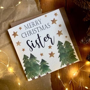 May include: A Christmas card with the message "Merry Christmas Sister" in black script and serif font. The card features watercolor illustrations of green pine trees and gold stars. A paper clip secures the card to a brown envelope.
