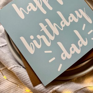 May include: A square greeting card with a light blue background and white text that reads happy birthday dad. The card is placed on top of a brown envelope, which is resting on a white plate. The design features simple, clean typography.