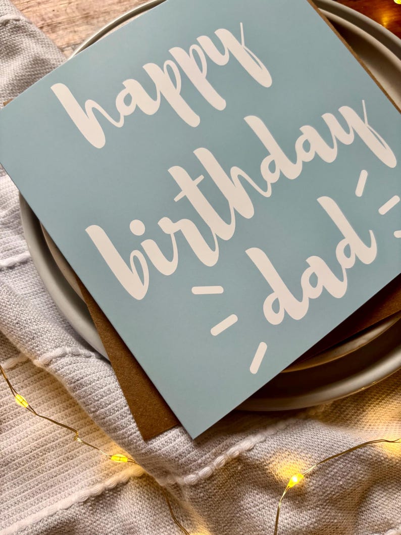May include: A square greeting card with a light blue background and white text that reads happy birthday dad. The card is placed on top of a brown envelope, which is resting on a white plate. The design features simple, clean typography.