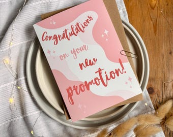 May include: A rectangular greeting card with a pink and white wavy background pattern. The text "Congratulations on your new promotion!" is printed in a dark red script font. Small white star icons are scattered across the card surface.