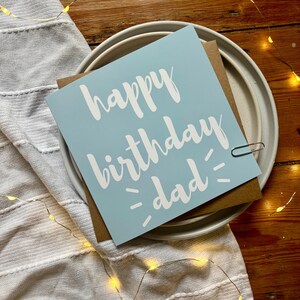 May include: A square greeting card with a light blue background and white text that reads happy birthday dad. The card is placed on a brown envelope, which rests on a white ceramic plate. A small silver paperclip is attached to the right side.
