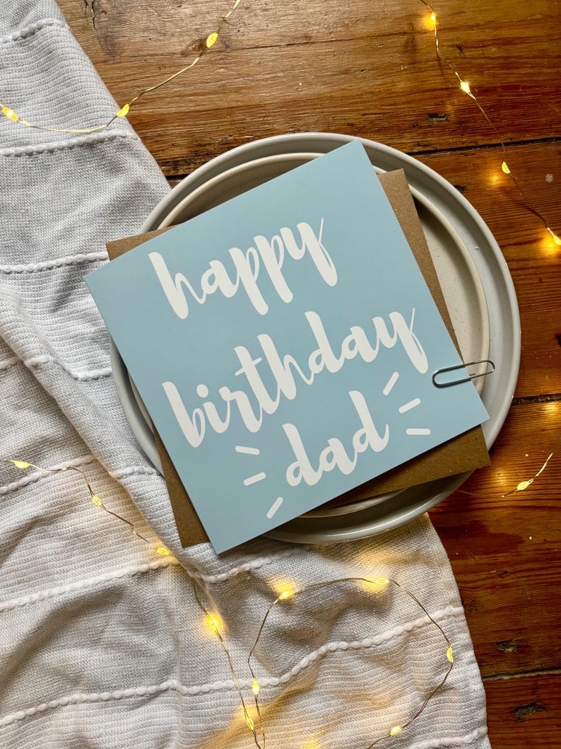 May include: A square greeting card with a light blue background and white text that reads happy birthday dad. The card is placed on a brown envelope, which rests on a white ceramic plate. A small silver paperclip is attached to the right side.