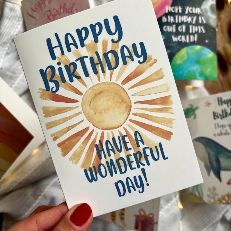 May include: A birthday card with the words "Happy Birthday" in navy blue script, and "Have a Wonderful Day!" below. The card features a watercolor sun design in shades of yellow and orange. Other cards are visible.