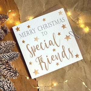 Puede incluir: Tarjeta de Navidad blanca con el mensaje "MERRY CHRISTMAS TO Special Friends" en letras marrones y decorada con estrellas marrones. Un clip plateado asegura la tarjeta. La tarjeta está sobre una superficie marrón, con piñas y luces.